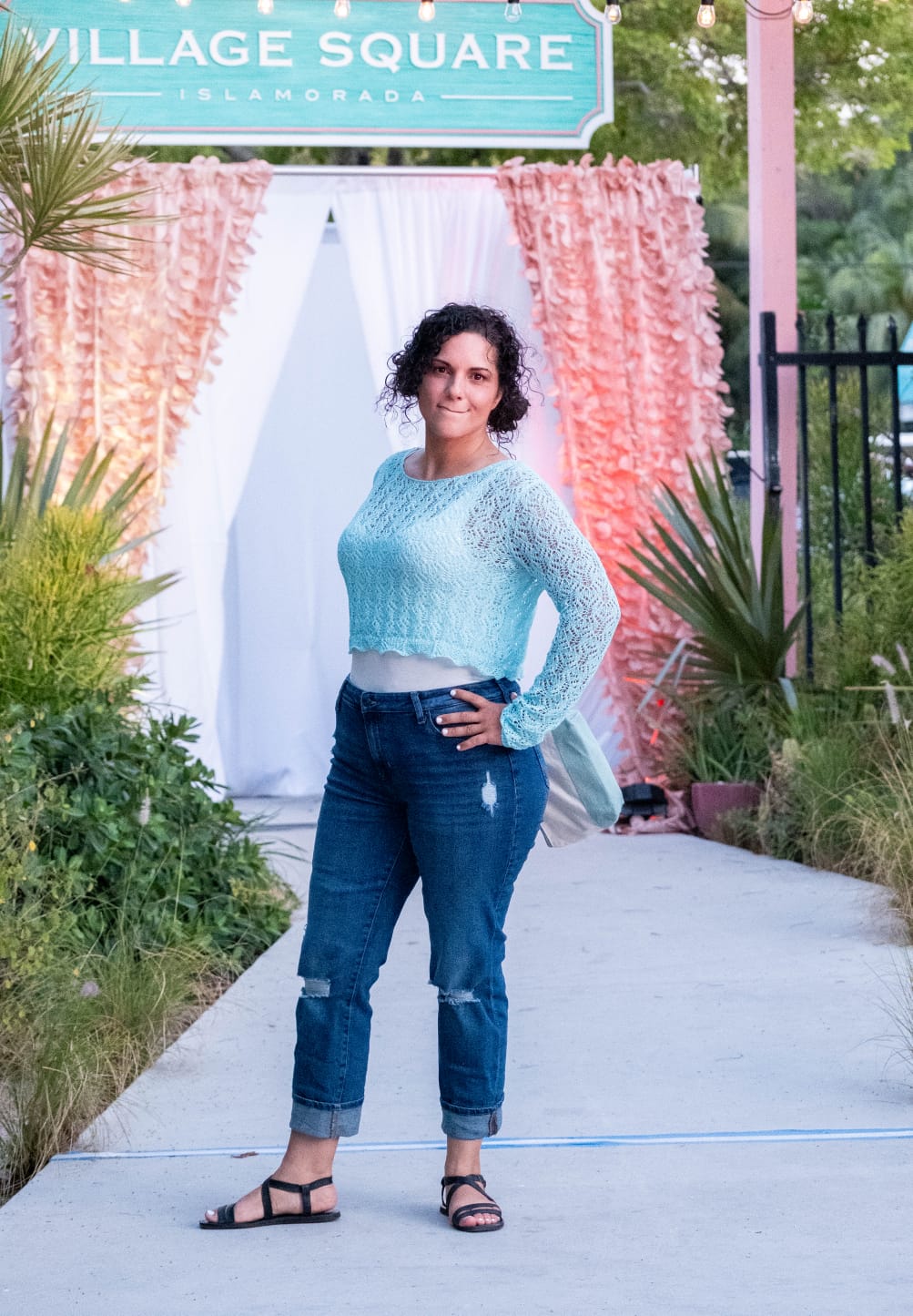 Woman in a light blue sweater and jeans standing in front of a decorative pink and white archway with 'Village Square' sign.
