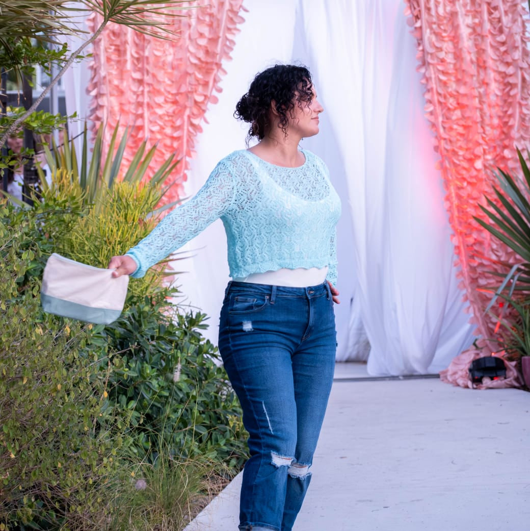 Woman in light blue sweater and jeans holding a clutch, with 'Village Square' sign in the background.